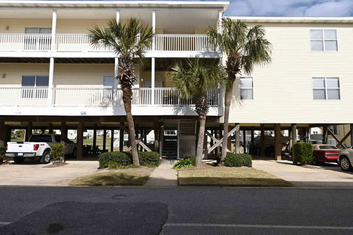 A two-story elevated building with private parking spaces underneath, a wooden stairway leading to the condo entrance, and palm trees in landscaped front. The setup is inviting, with a clear view of the parking and entry area.