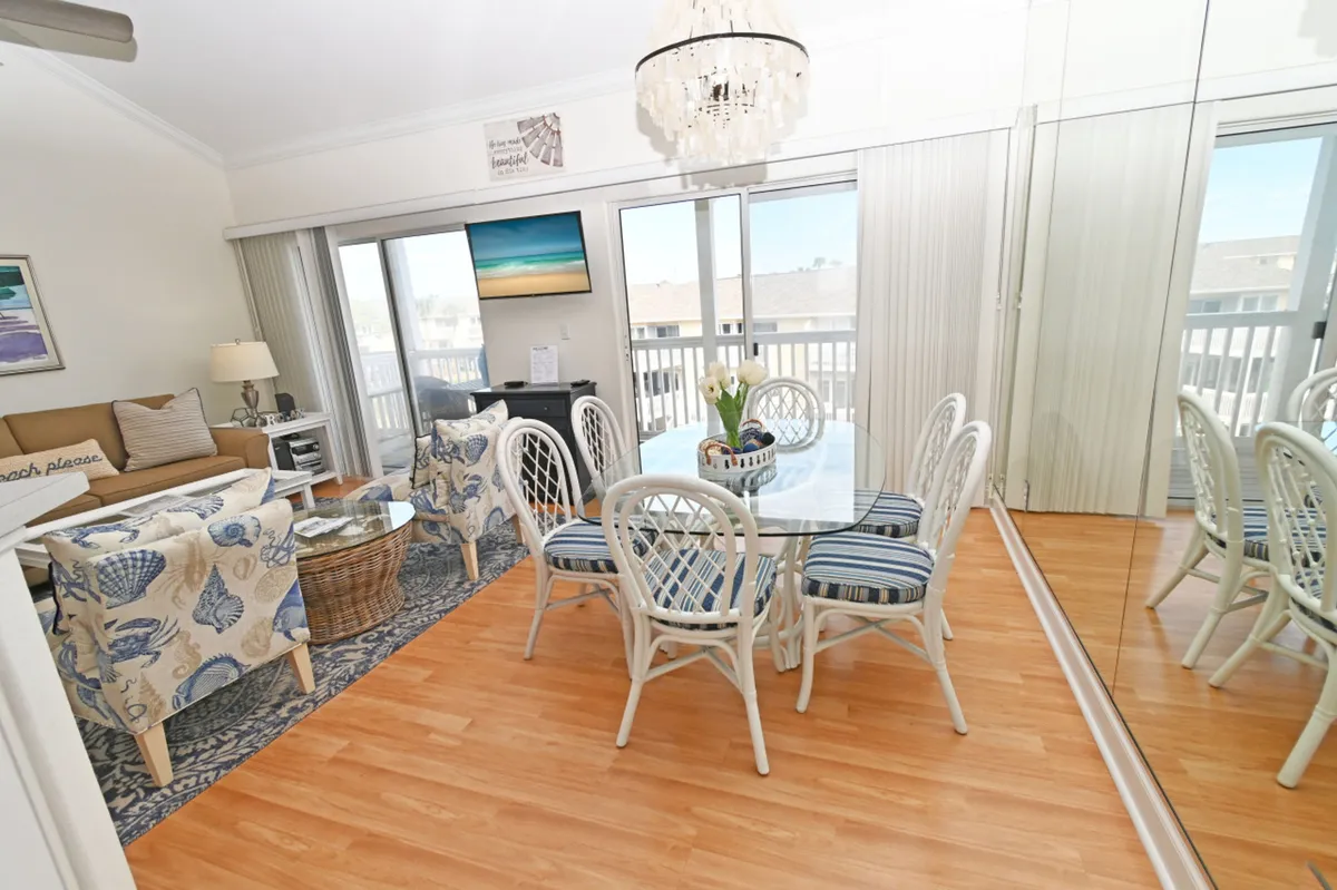 A bright, cheerful dining and living area features a glass dining table with striped cushioned chairs, a cozy seating area with seashell-patterned armchairs, a mirrored wall reflecting abundant natural light, and sliding glass doors leading directly to the private balcony.
