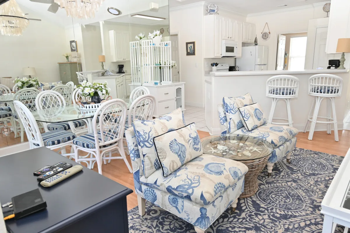 A cheerful, coastal-inspired open-plan living and dining area with white kitchen cabinetry, a breakfast bar with stools, a dining table with rattan chairs, and cozy seating with beachy decor. Natural light fills the space, creating an inviting atmosphere for family gatherings and casual dining.