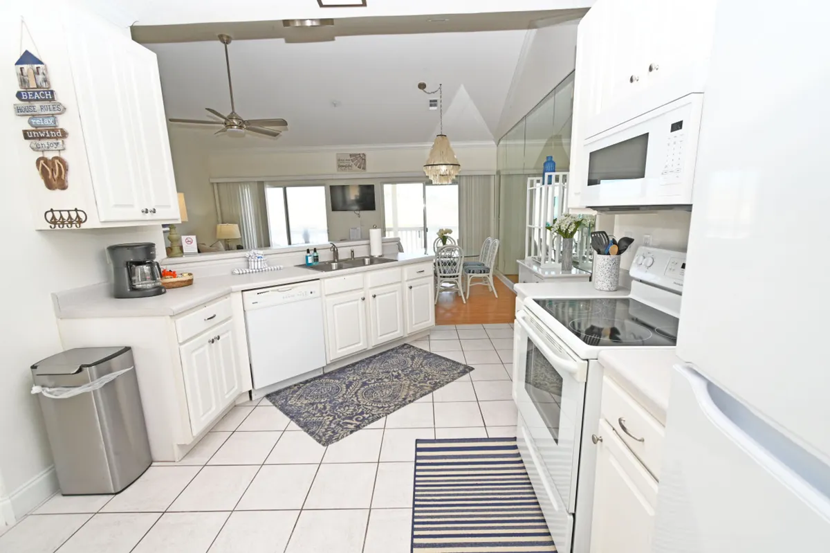 A bright, well-lit kitchen featuring white cabinetry and countertops, modern stainless appliances like a microwave and stove, a decorative beach-themed sign, and a dining area visible through a glass wall. The breakfast bar and open layout invite casual dining and social interaction.