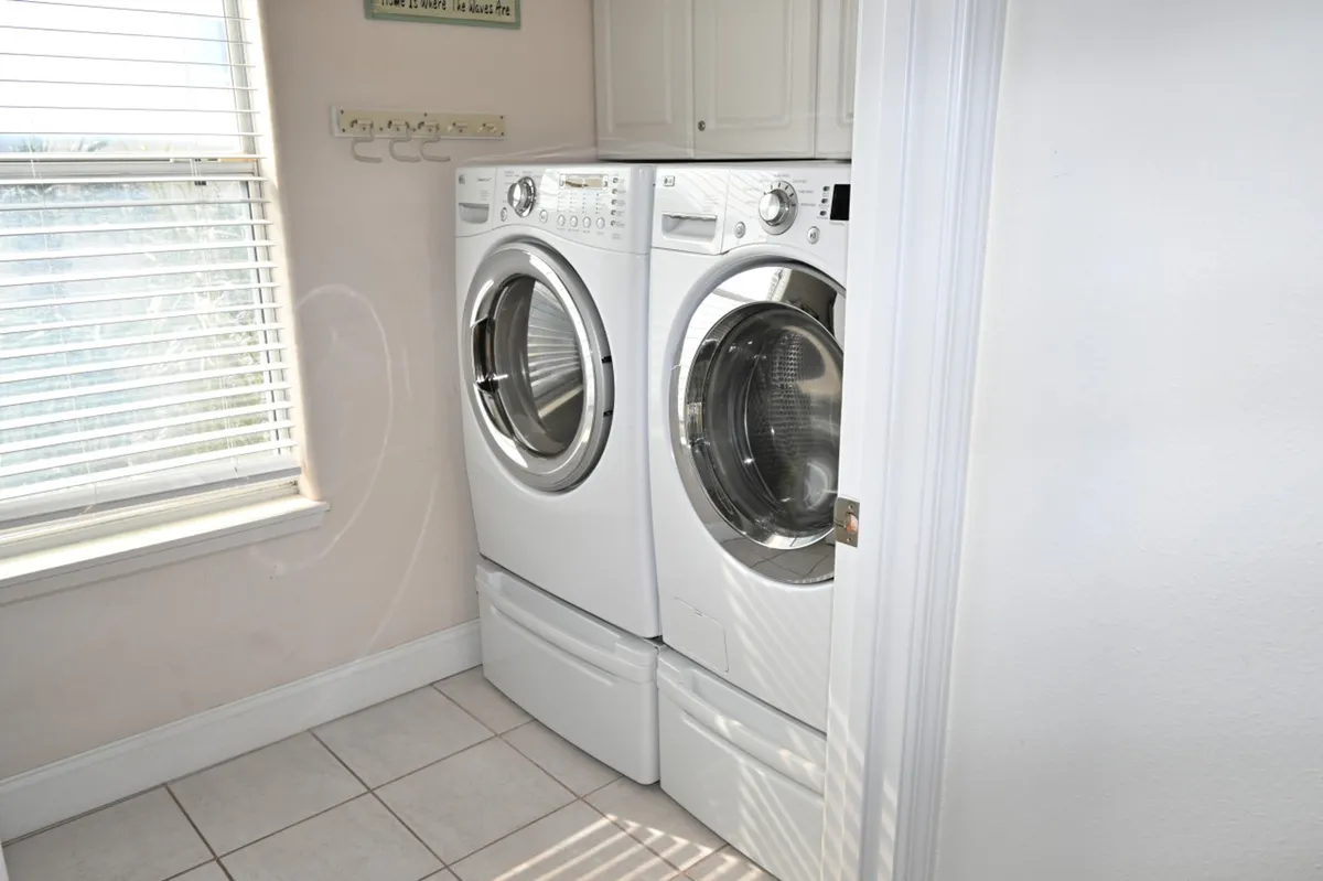 A bright, well-lit laundry room featuring a front-loading washing machine and dryer side by side, under white cabinetry. Natural light pours in through a window with blinds, and the space feels clean and organized.