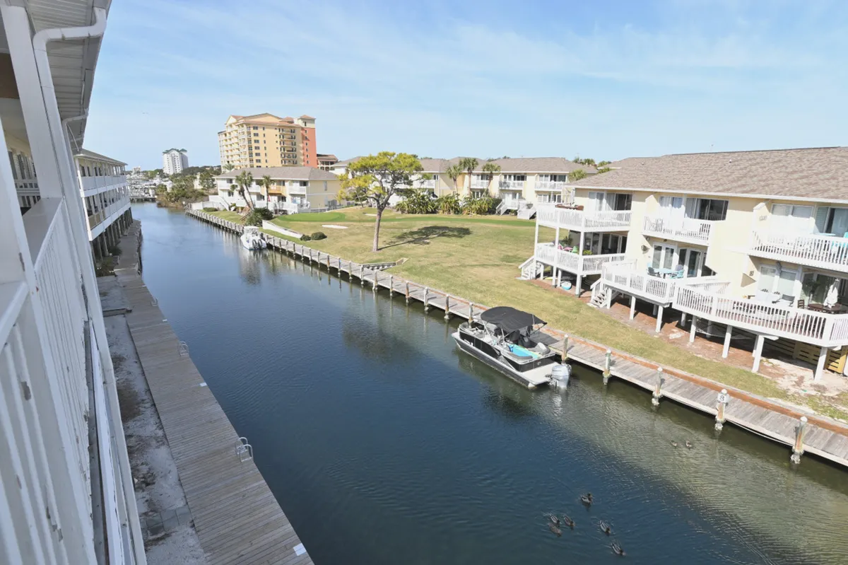 A canal scene with a multi-level residential building featuring balconies, a grassy communal lawn area, and ducks swimming in the canal. The pet-friendly, open lawn and water access are prominent.