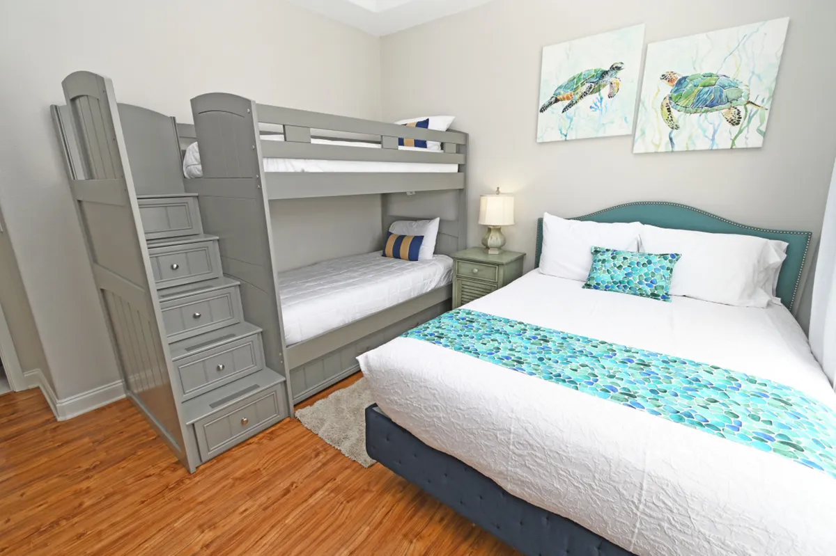 The image displays a guest bedroom with a queen-size bed adorned in blue and turquoise accents, a grey bunk bed with built-in staircase drawers, and playful sea turtle paintings on the wall. There's also a green nightstand with a lamp, creating a cozy, family-friendly environment.