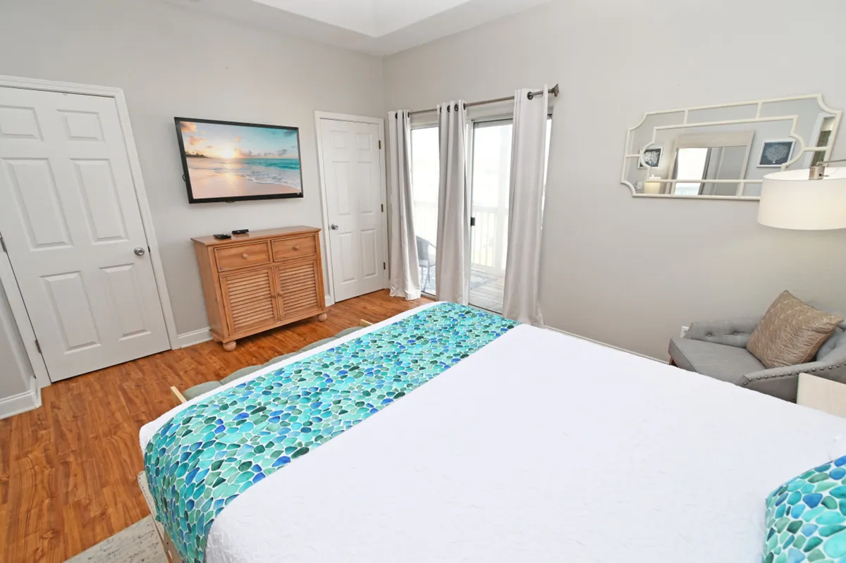 This image showcases a spacious bedroom featuring a large bed with blue and green patterned bedding, a wall-mounted flat-screen TV displaying a beach scene, a wooden dresser, and a sliding glass door leading to a balcony. The room is well-lit and inviting, emphasizing comfort and a private retreat atmosphere.