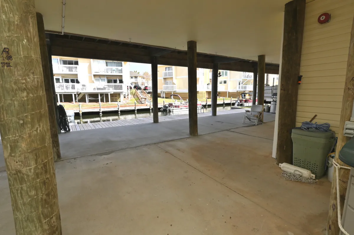 A practical, covered parking space beneath the unit with a view of docked boats on the canal, adjacent residential buildings, and some storage and seating items at the side. The concrete floor and wooden support pillars emphasize utility and accessibility.