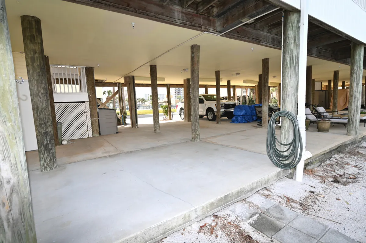 A practical view of the private parking space located underneath the condo, featuring wooden support pillars, parked vehicles including a white truck, and a coiled garden hose. The area is utilitarian and designed for easy access and unloading.