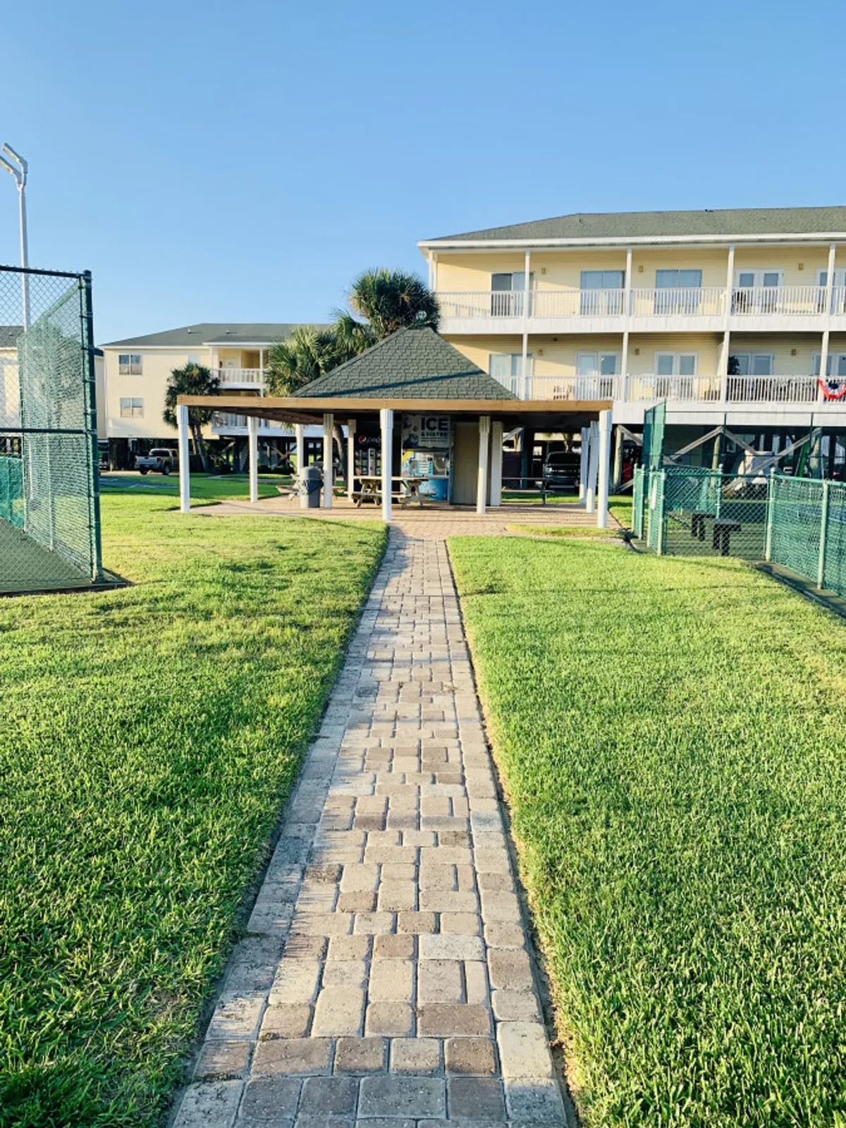 An inviting paved walkway leads to a gazebo structure and a covered pavilion with picnic tables and vending machines. Tennis or pickleball courts are fenced on both sides, and a three-story condominium building is visible in the background. The green space and winding path evoke a sense of a well-kept, active community.