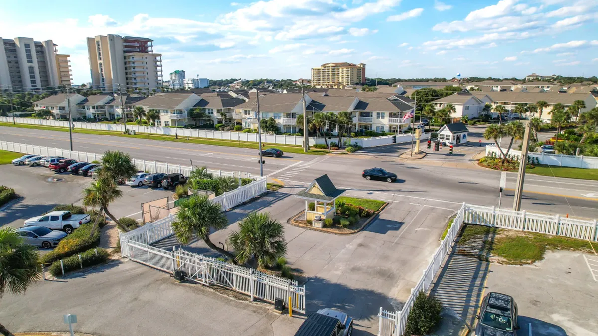 The gated entrance to Sandpiper Cove is shown, complete with a white security fence, a small guard house, and visible parking spaces. Distant residential buildings and condos are seen beyond the entrance, suggesting a secure, welcoming community.