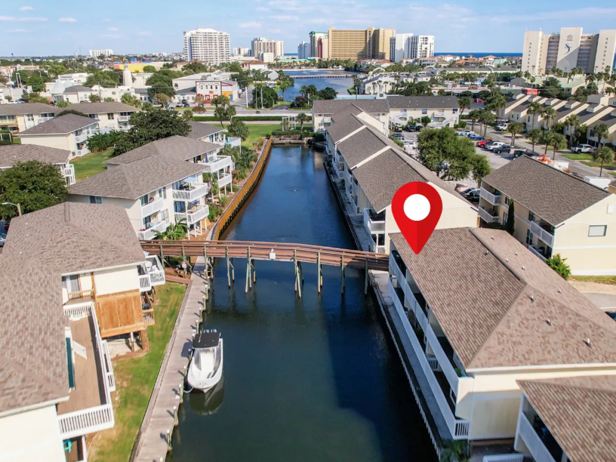This exterior shot showcases a private canal winding through the Sandpiper Cove community, with a docked boat, a bridge over the canal, and low-rise condo buildings with balconies. The Gulf of Mexico and high-rise hotels can be seen in the distance. The pathway alongside the canal is visible, inviting leisurely walks and exploration.