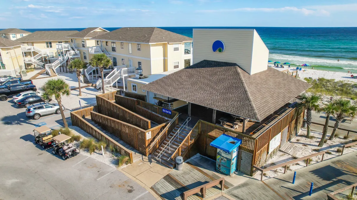 This exterior image features a beachfront restaurant or bar with shaded patios, umbrellas dotting the sandy beach, waves in the background, and a lively vacation ambiance. Low-rise buildings and a parking lot are visible, reinforcing the resort setting.