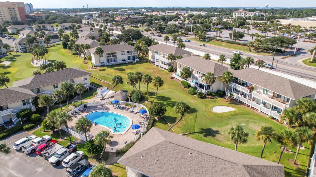 A lively exterior scene of the community swimming pool with people enjoying the water, surrounded by lush palm trees, condo buildings, and a visible golf course green with a sand trap. The setting radiates a resort-like, vacation mood and social atmosphere.