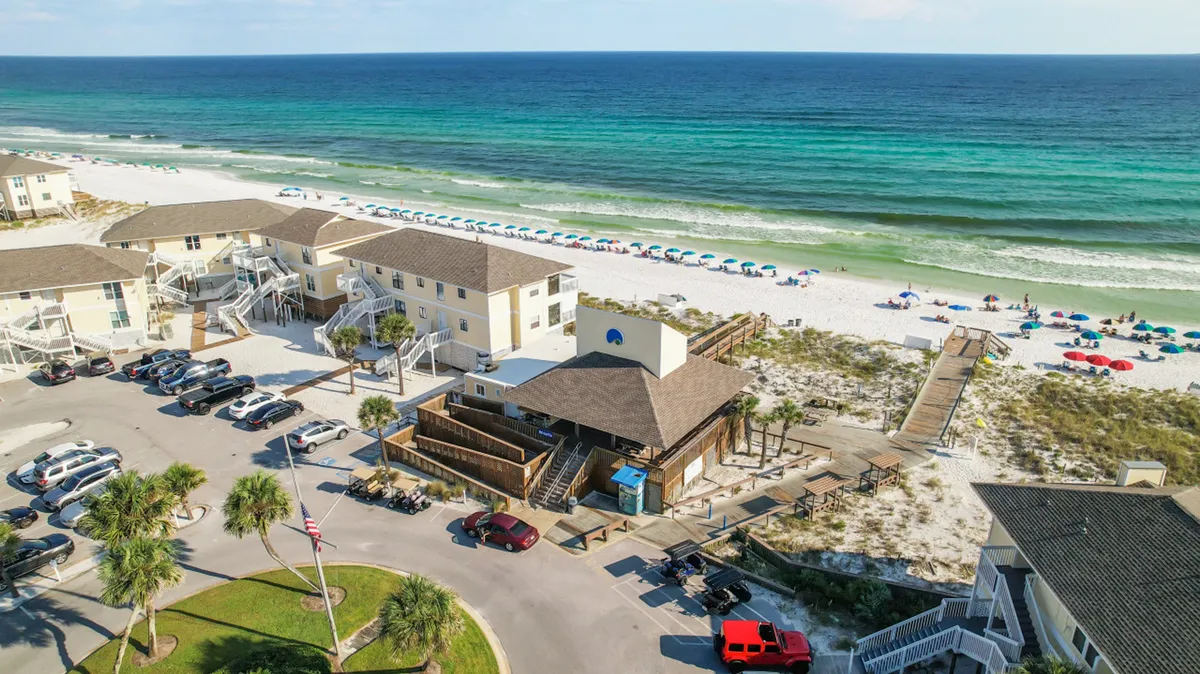 This vibrant exterior shot captures a white sandy beach with colorful umbrellas dotting the shoreline, turquoise ocean waves, a beach access boardwalk, and nearby resort buildings. People are seen lounging and enjoying the beach, evoking the quintessential beach day experience.
